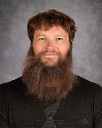 Portrait of a man with brown hair and a long brown beard, wearing a dark shirt against a gray backdrop
