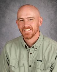 Portrait of a bald man with a red beard, smiling in a light green button-up shirt against a gray backdrop.