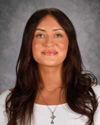 Portrait of a person with long dark hair, wearing a white top and necklace against a gray backdrop