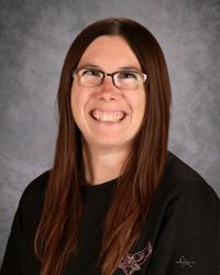 Smiling person with glasses and long brown hair against a gray studio backdrop