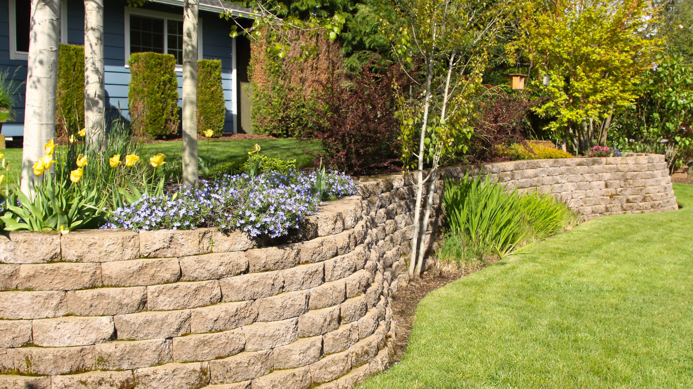 Stone retaining walls with flowers and bushes in front of a light blue house and green lawn.