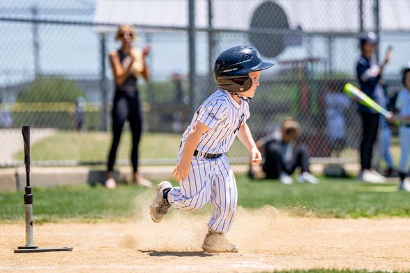 Child running on a baseball field toward a base, kicking up dust, with spectators in the background