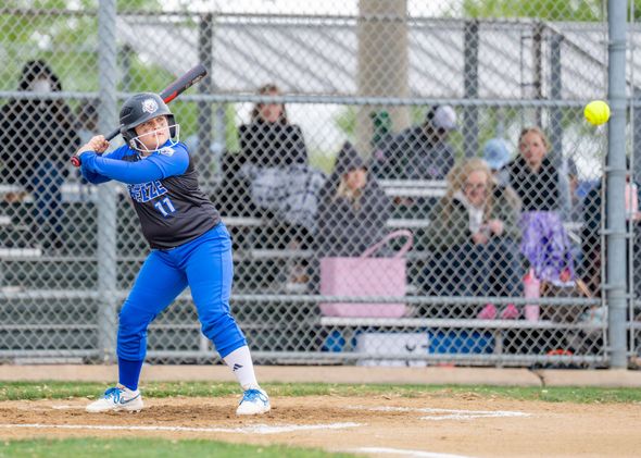Softball player in blue uniform swings at a pitch on a field, with spectators behind a chain-link fence