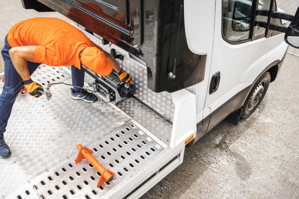 A worker in an orange shirt and hat working on the back of a white tow truck, using a tool.