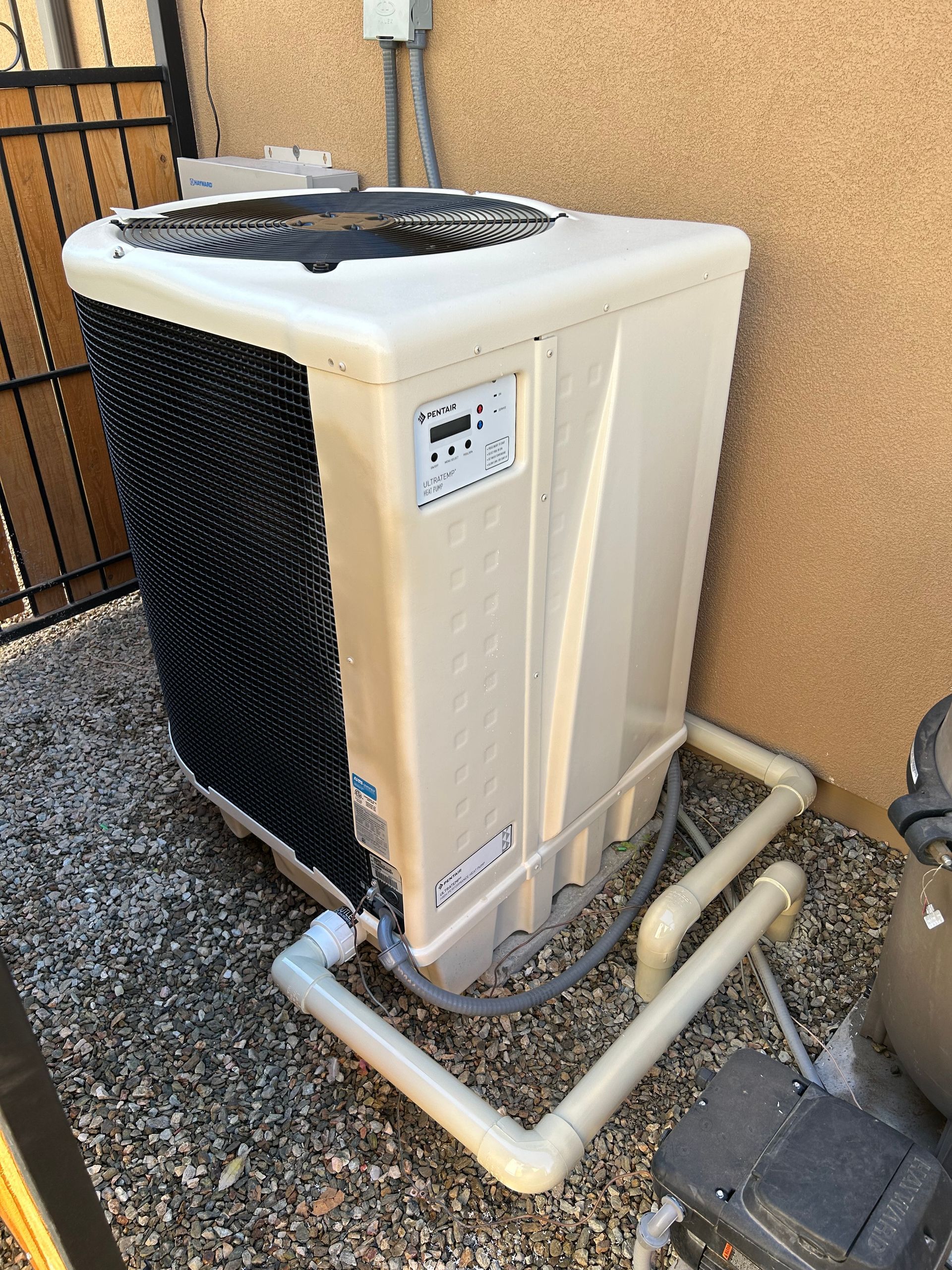 Pool heat pump near a wall and fence, with PVC pipes and a digital control panel.
