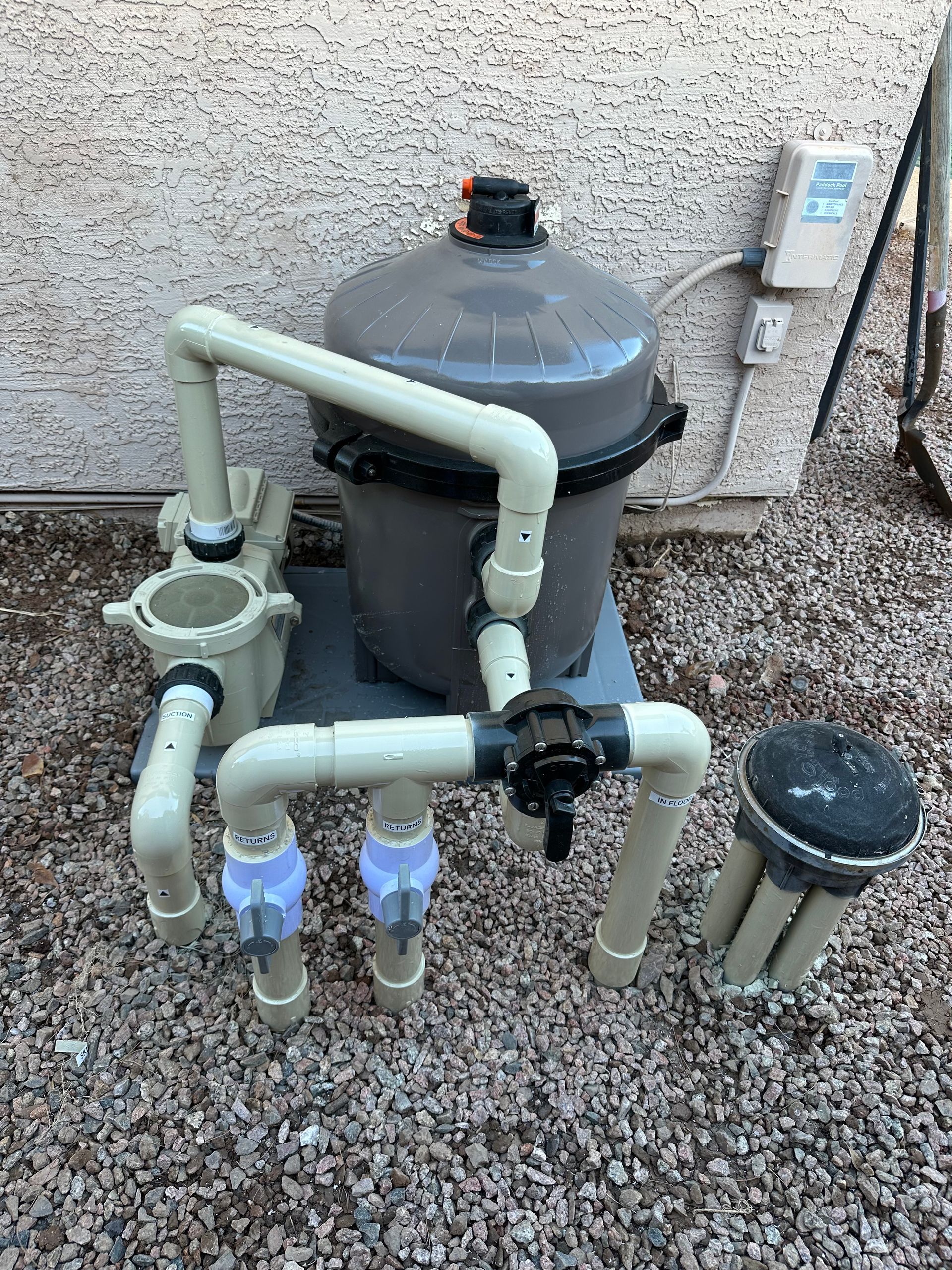 Pool equipment: pump, filter, and plumbing on gravel next to a wall.