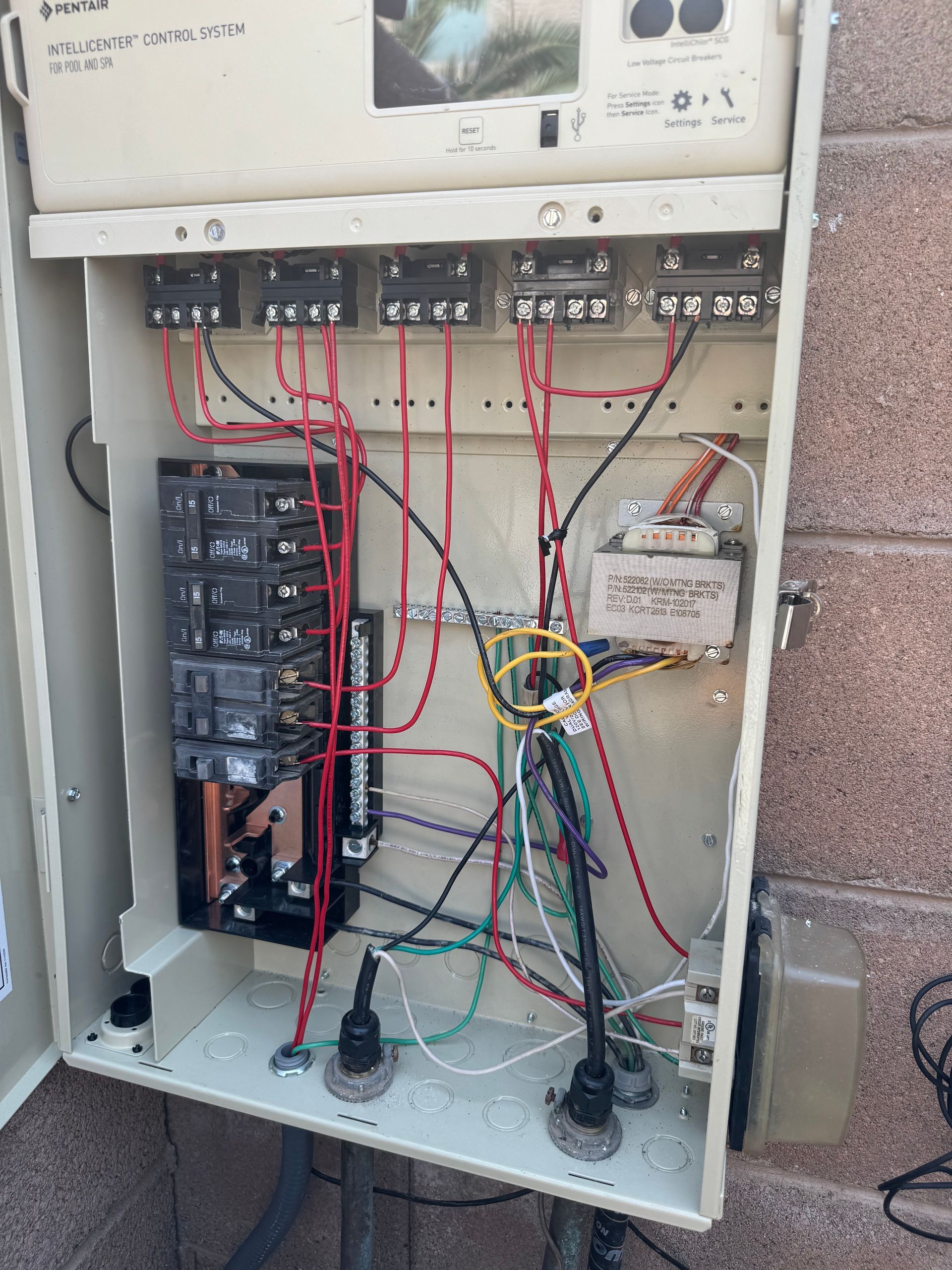 Open beige electrical box with complex wiring, circuit breakers, and transformer against a brick wall.