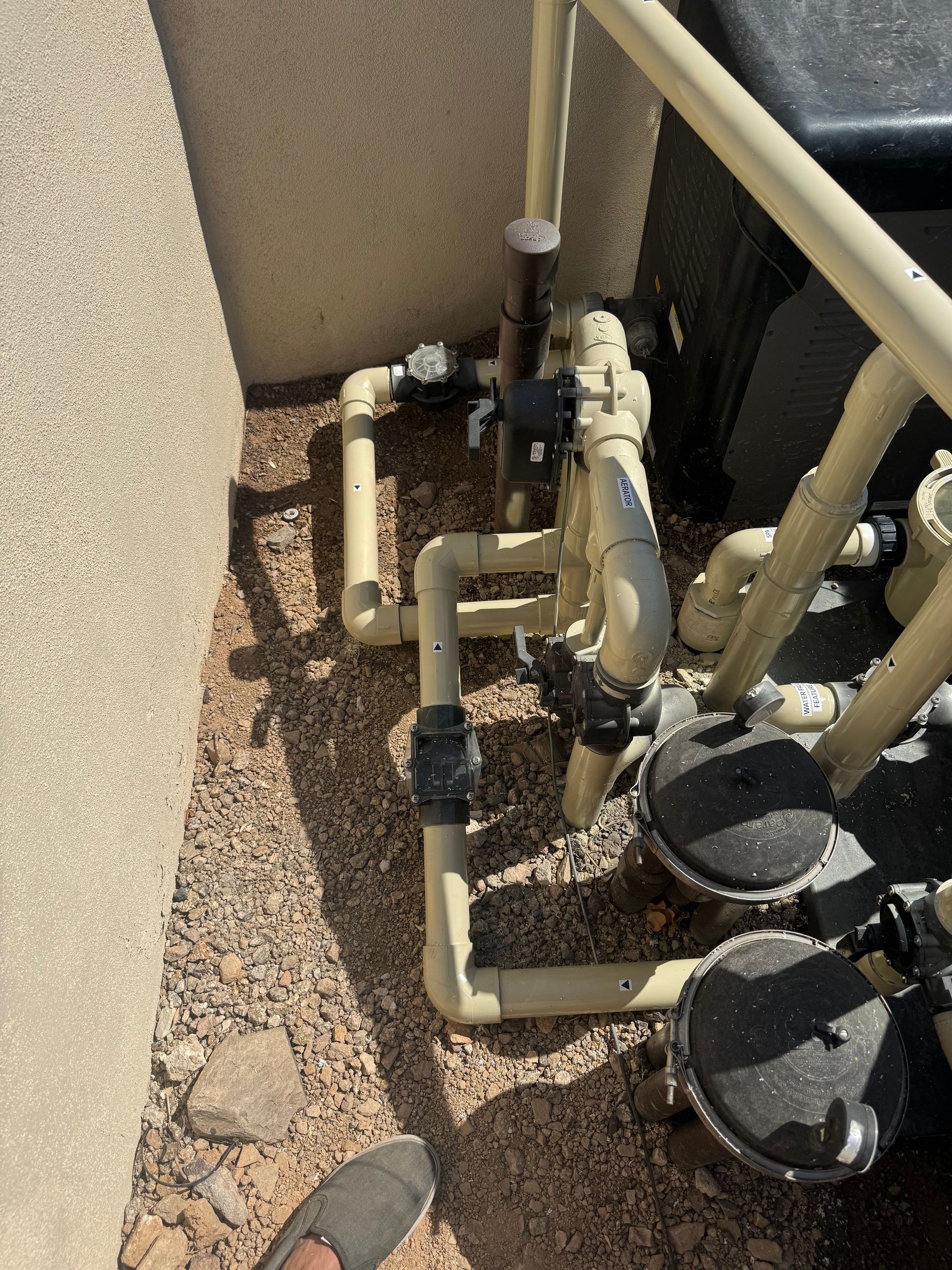 Pool equipment with tan pipes, black valves, and filter components against a stucco wall.