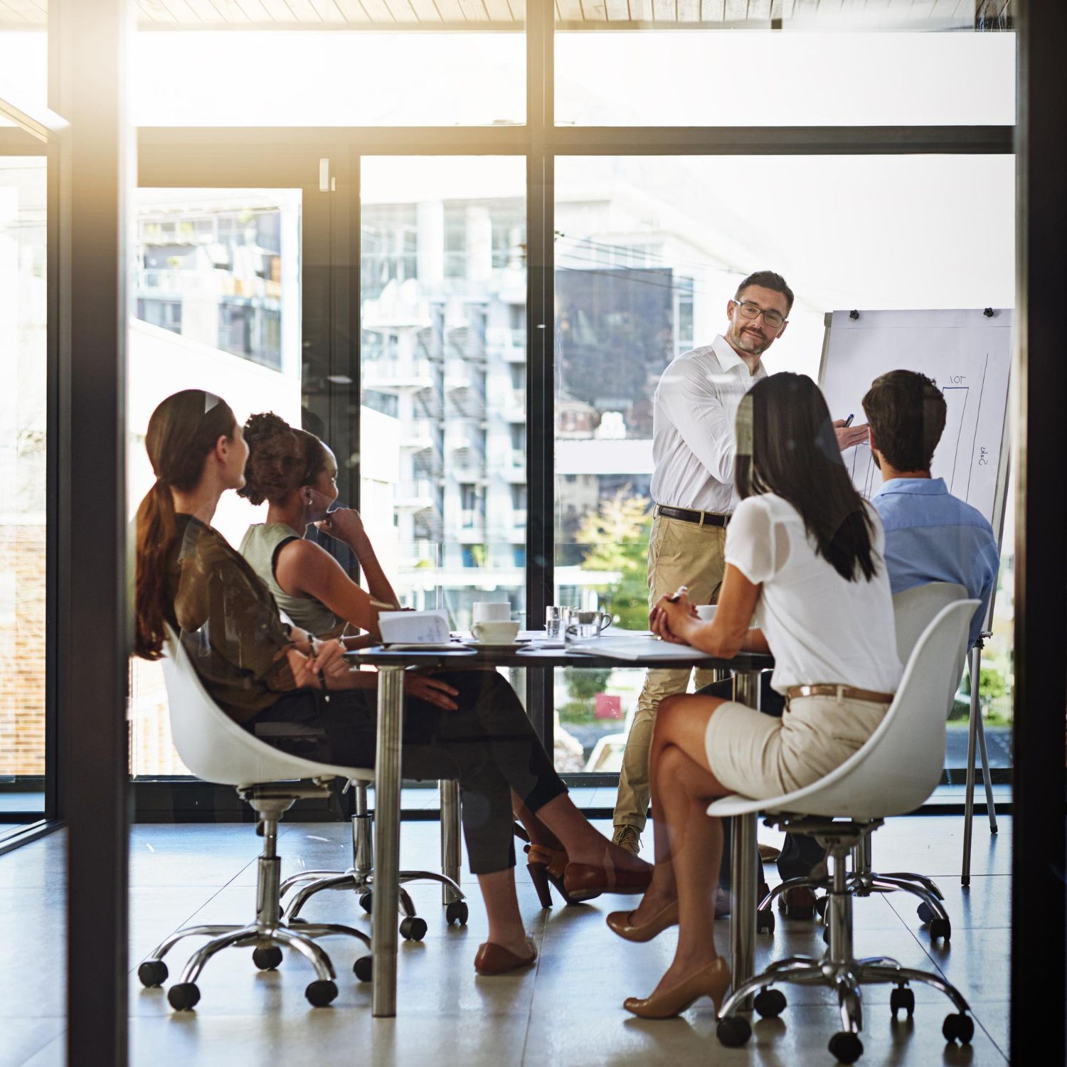 Business team meeting around a table, one person presenting at a whiteboard in a bright office.