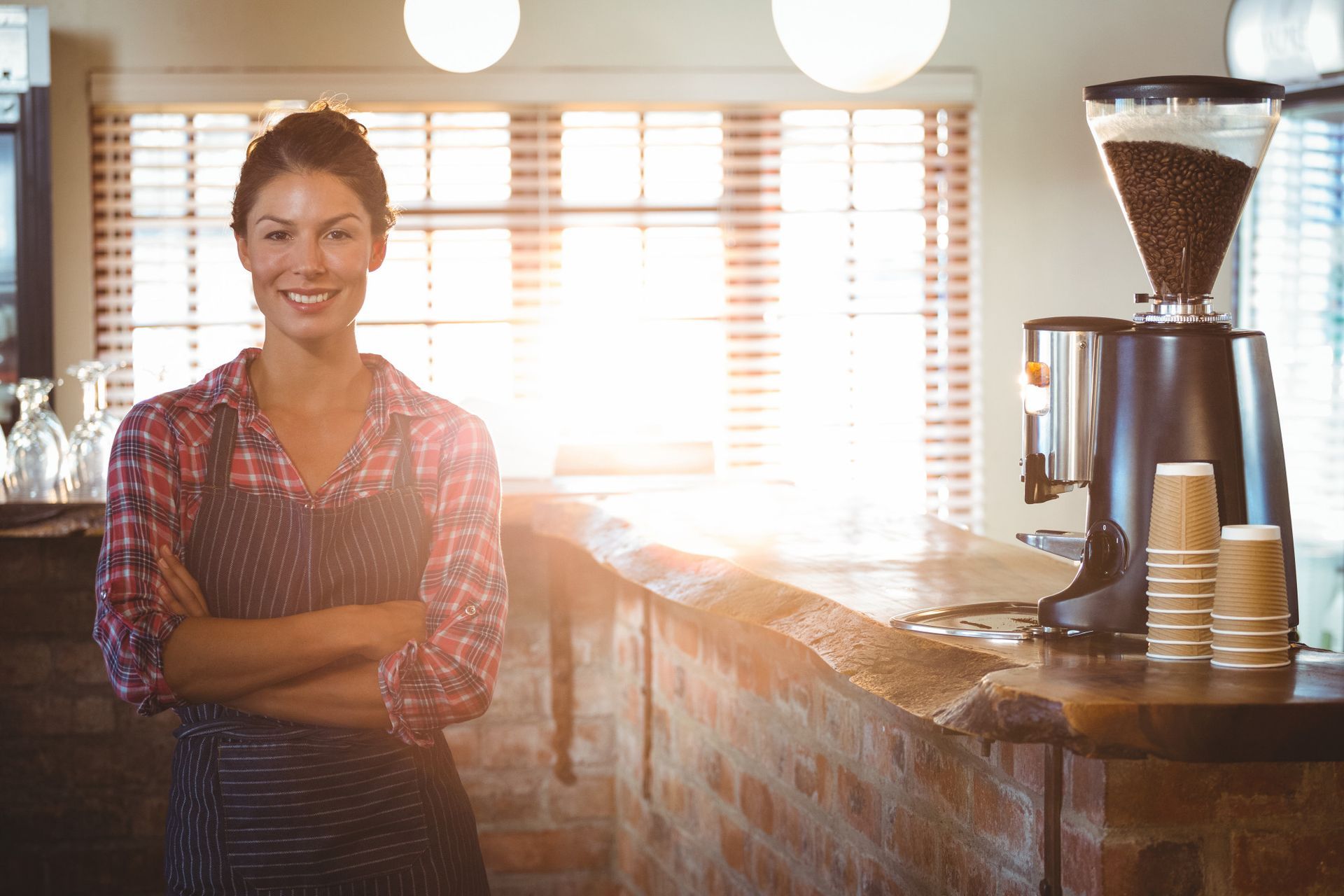 Woman in apron, arms crossed, smiling at coffee shop counter with coffee grinder and cups.