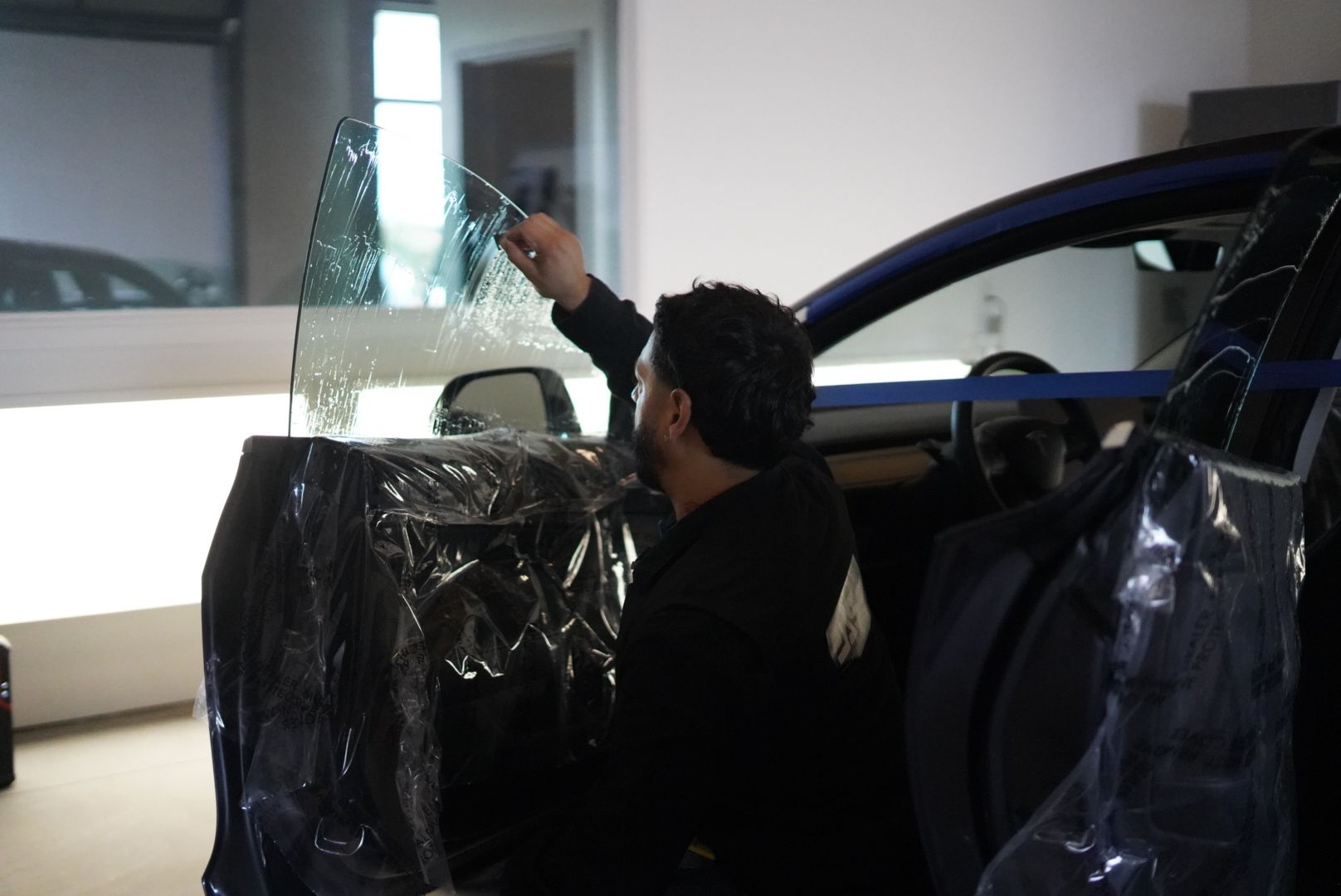 Man installing a car windshield in a workshop, with a blue vehicle door open nearby