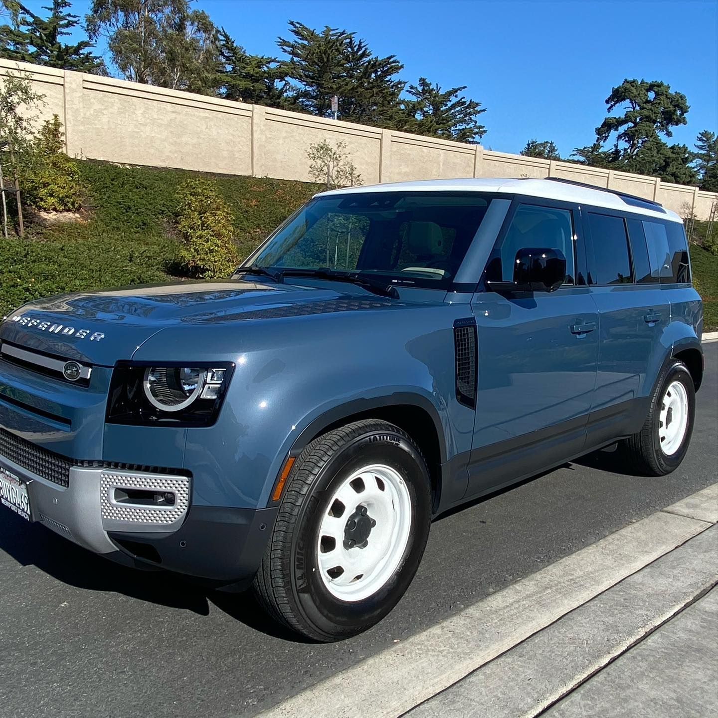 Blue Land Rover Defender parked on a sunny street beside a hedge and beige wall