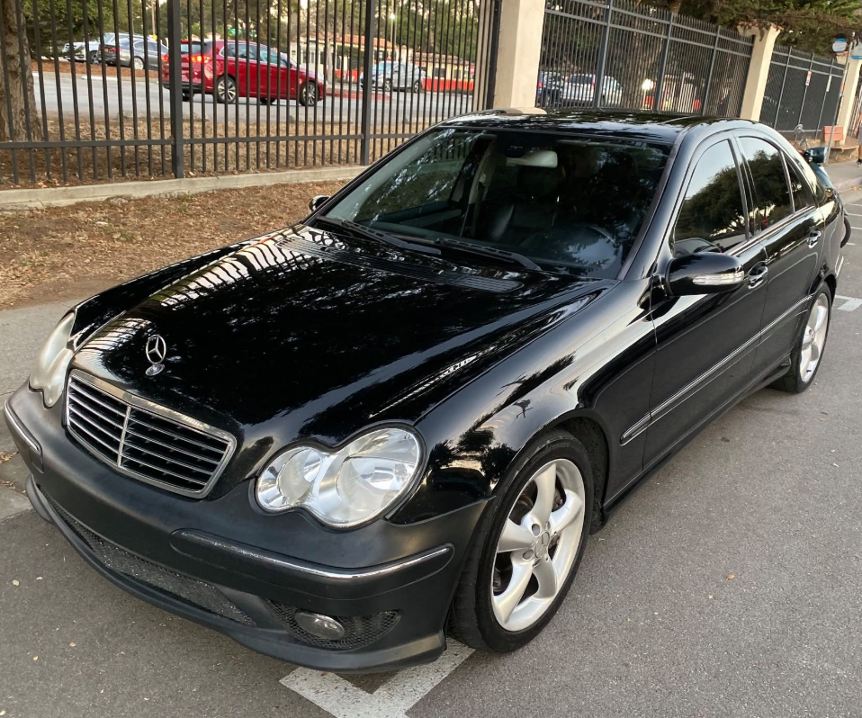 Black Mercedes sedan parked on a street beside a fence, viewed from the front-left angle