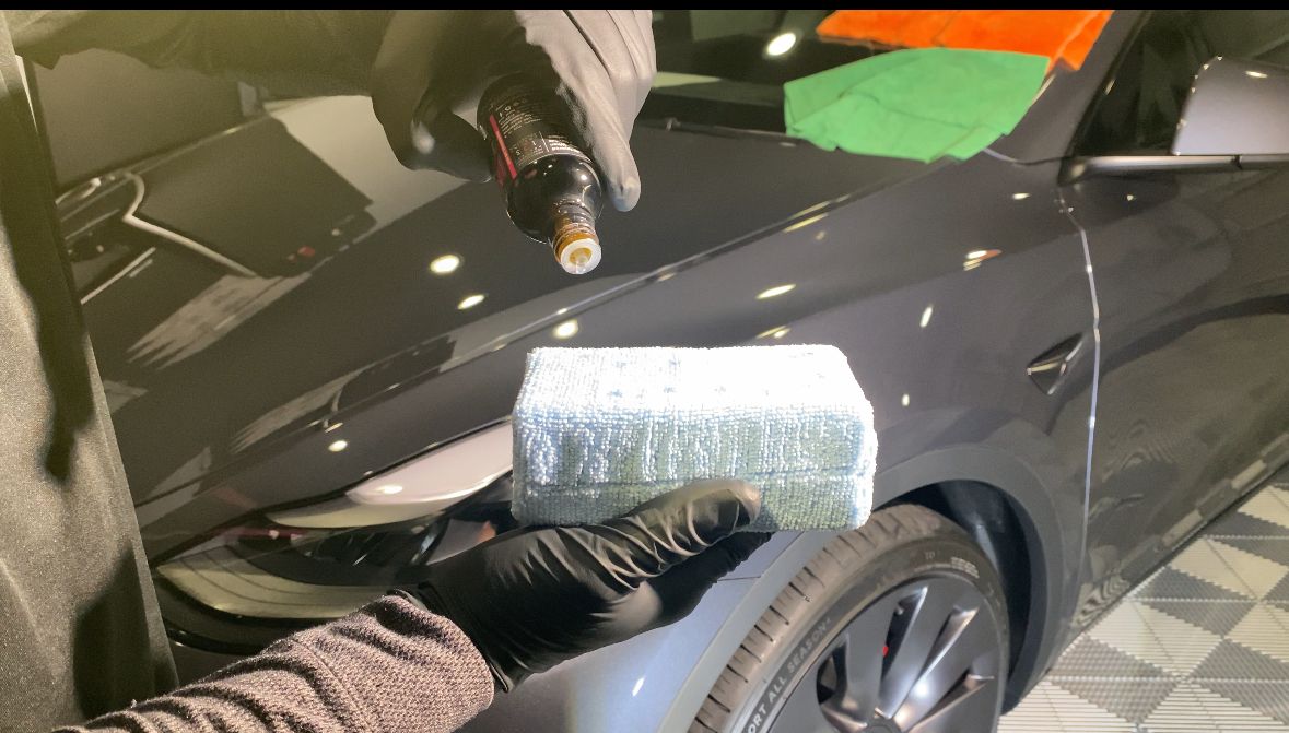 Person polishing a black car hood with a sponge and spray bottle in a garage