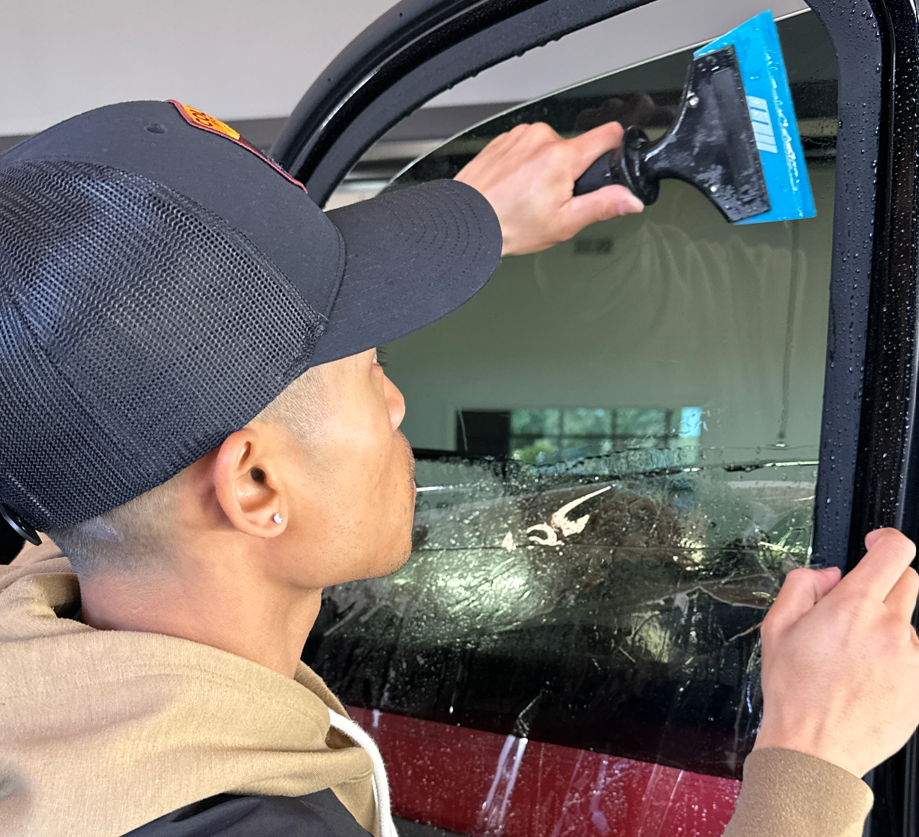Person cleaning a car window with a blue squeegee, wearing a black cap and tan hoodie