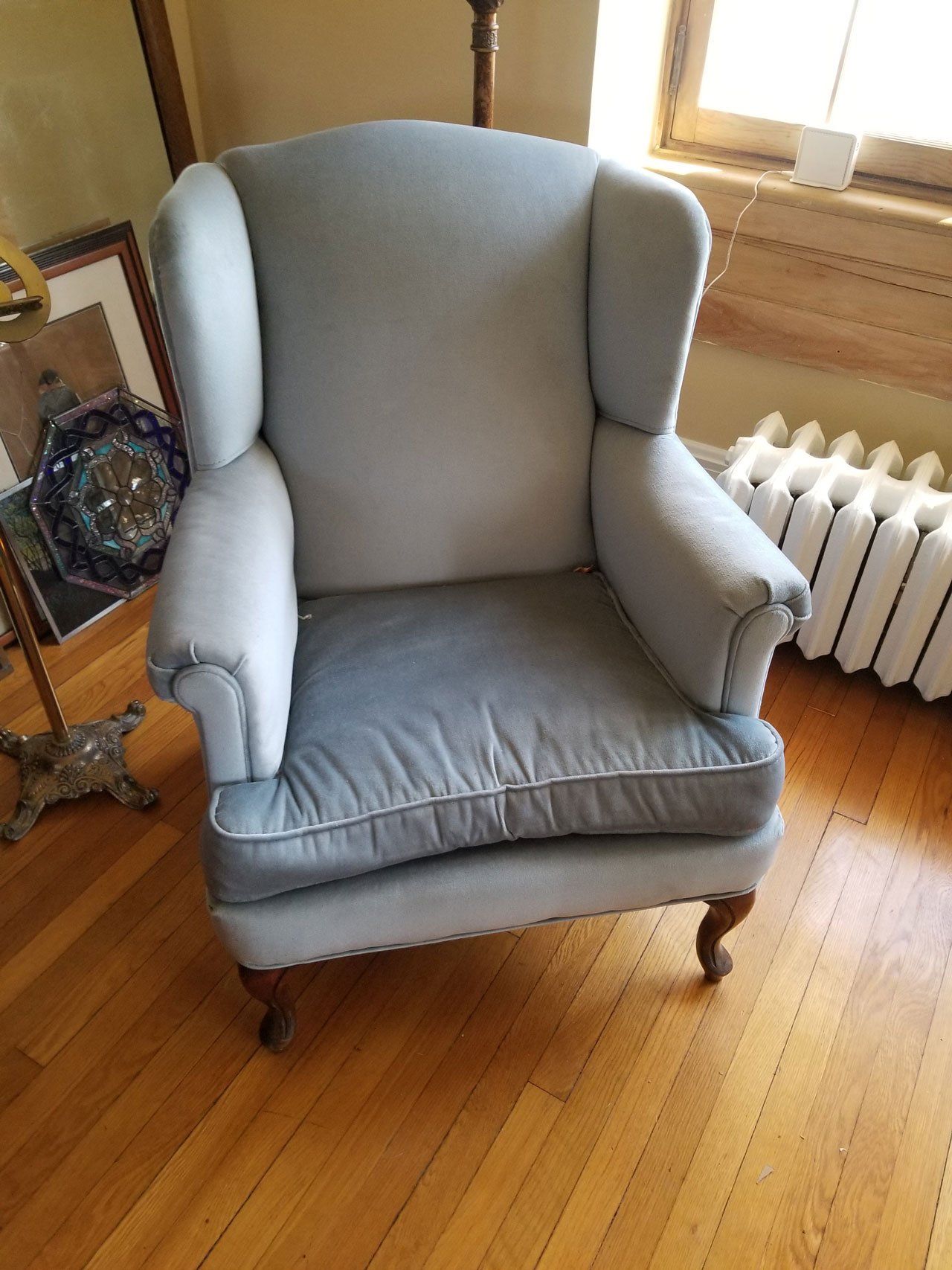 A light blue upholstered wingback chair with wooden legs, positioned on a wood floor near a white radiator.