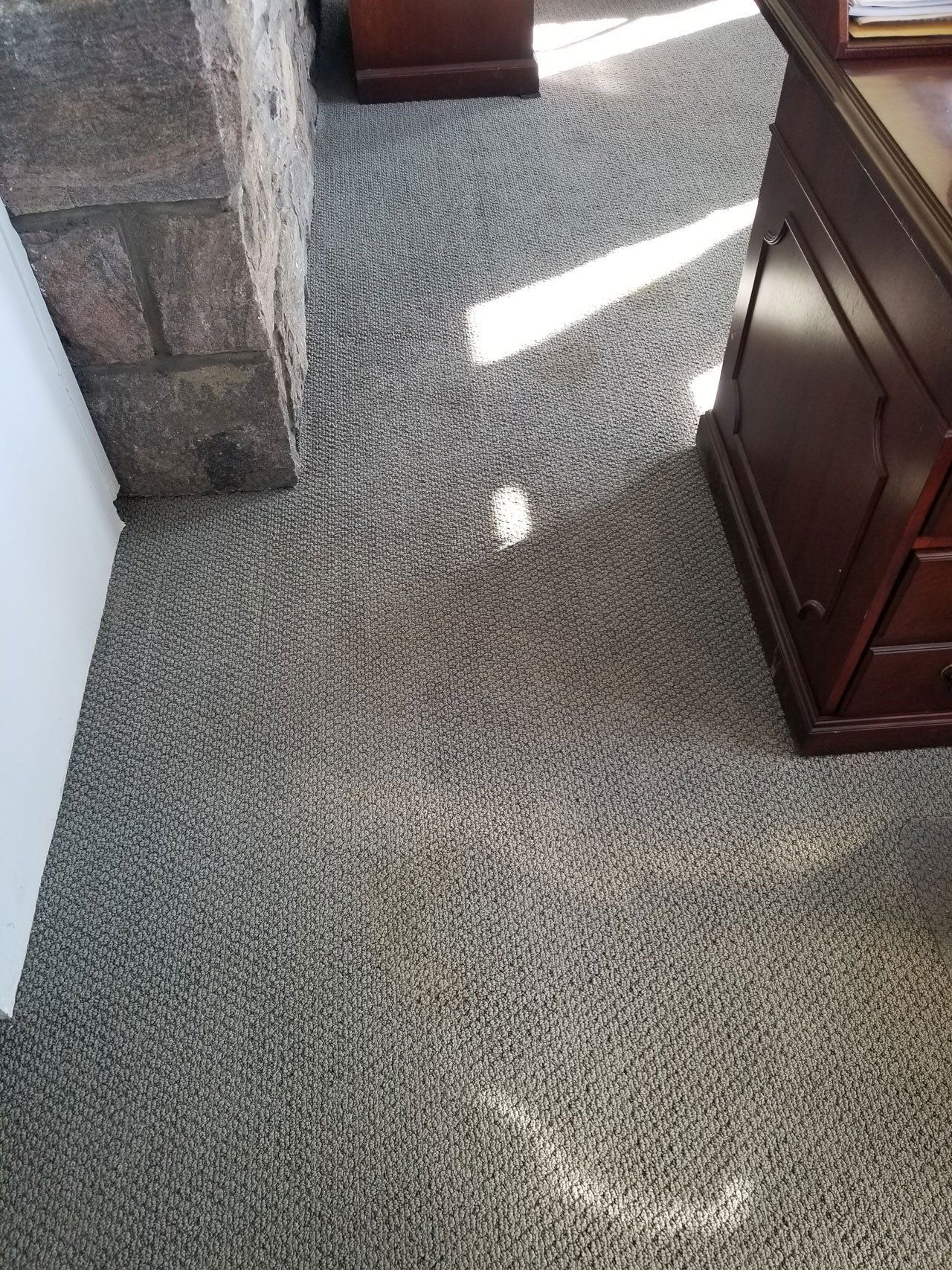 A high-angle view of gray, speckled office carpet between a stone pillar and a wooden desk base in a bright room.