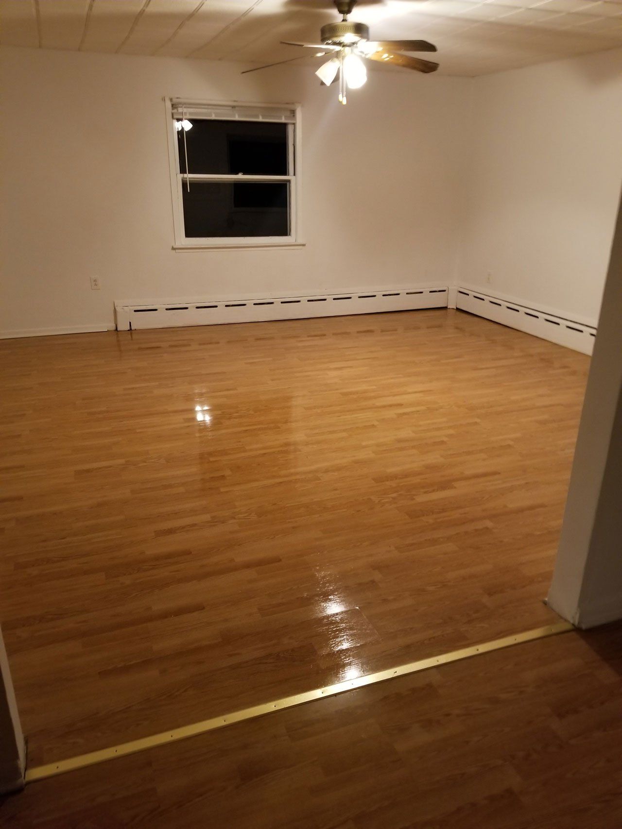 An empty room with glossy, wood-look laminate flooring, white walls, and a ceiling fan over a single window at night.