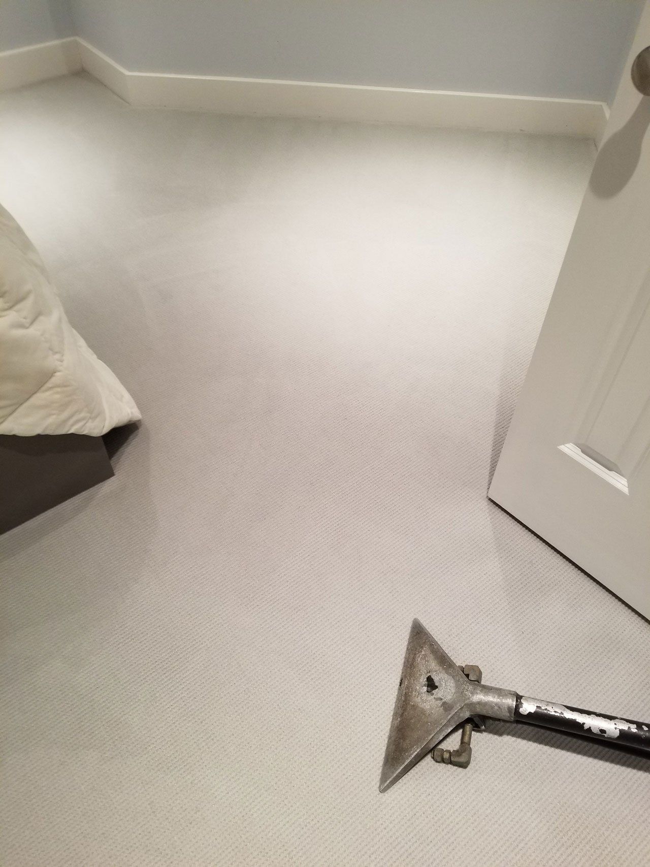 A carpet cleaning wand sits on light-colored carpet in a bedroom with a partial view of a bed and a white door.