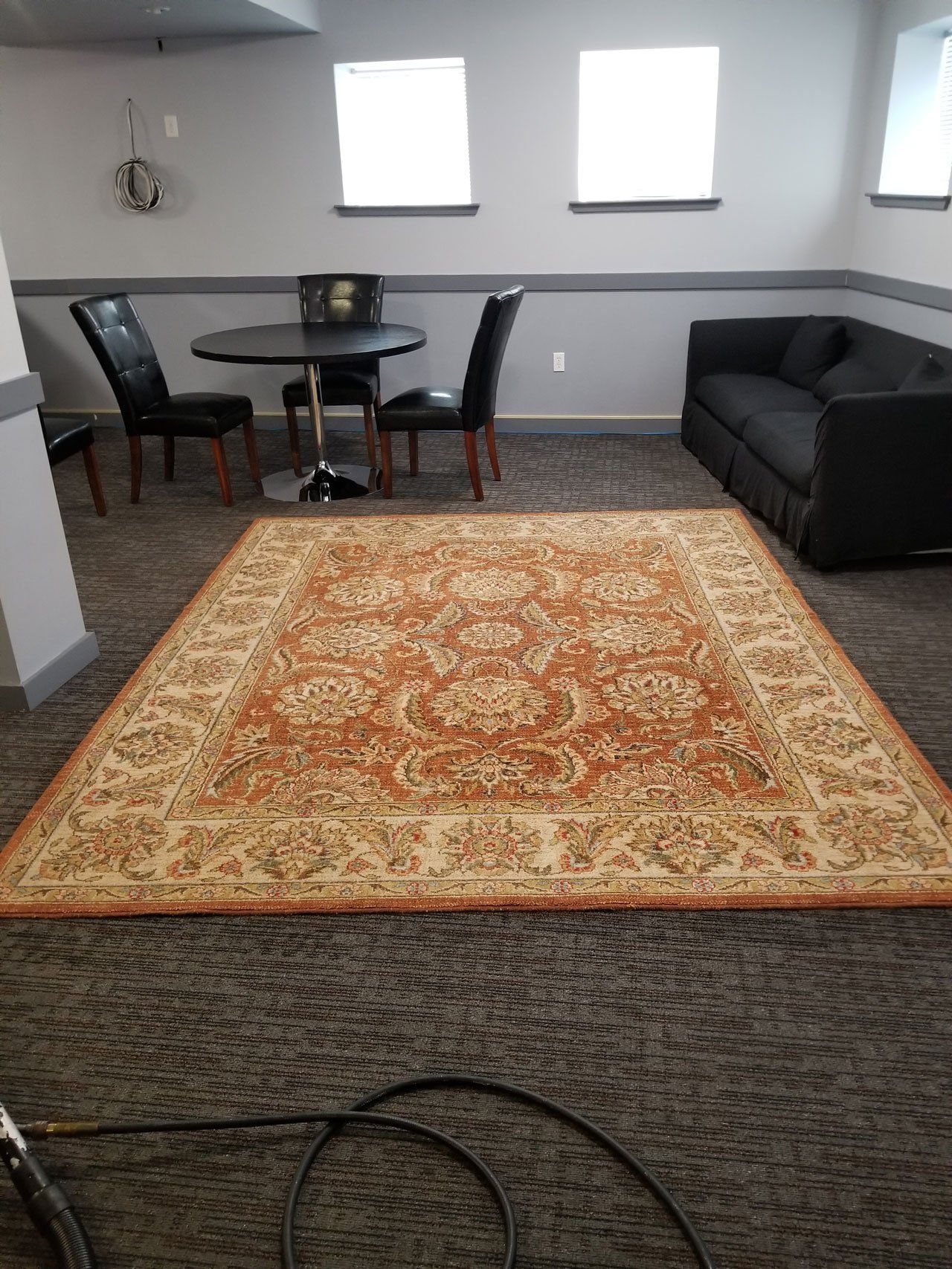 A room with gray carpet, a table with three black chairs, a black sofa, and a large, ornate orange and cream area rug.