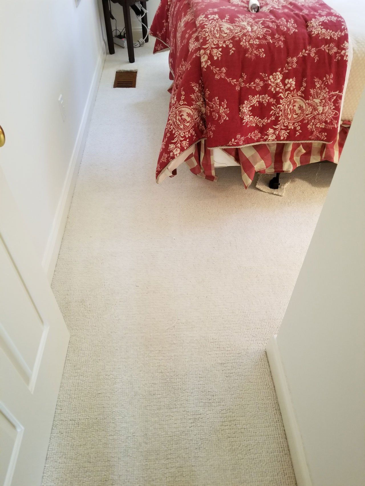 An indoor shot of a beige carpeted floor, leading toward a bed with a red floral patterned bedspread.