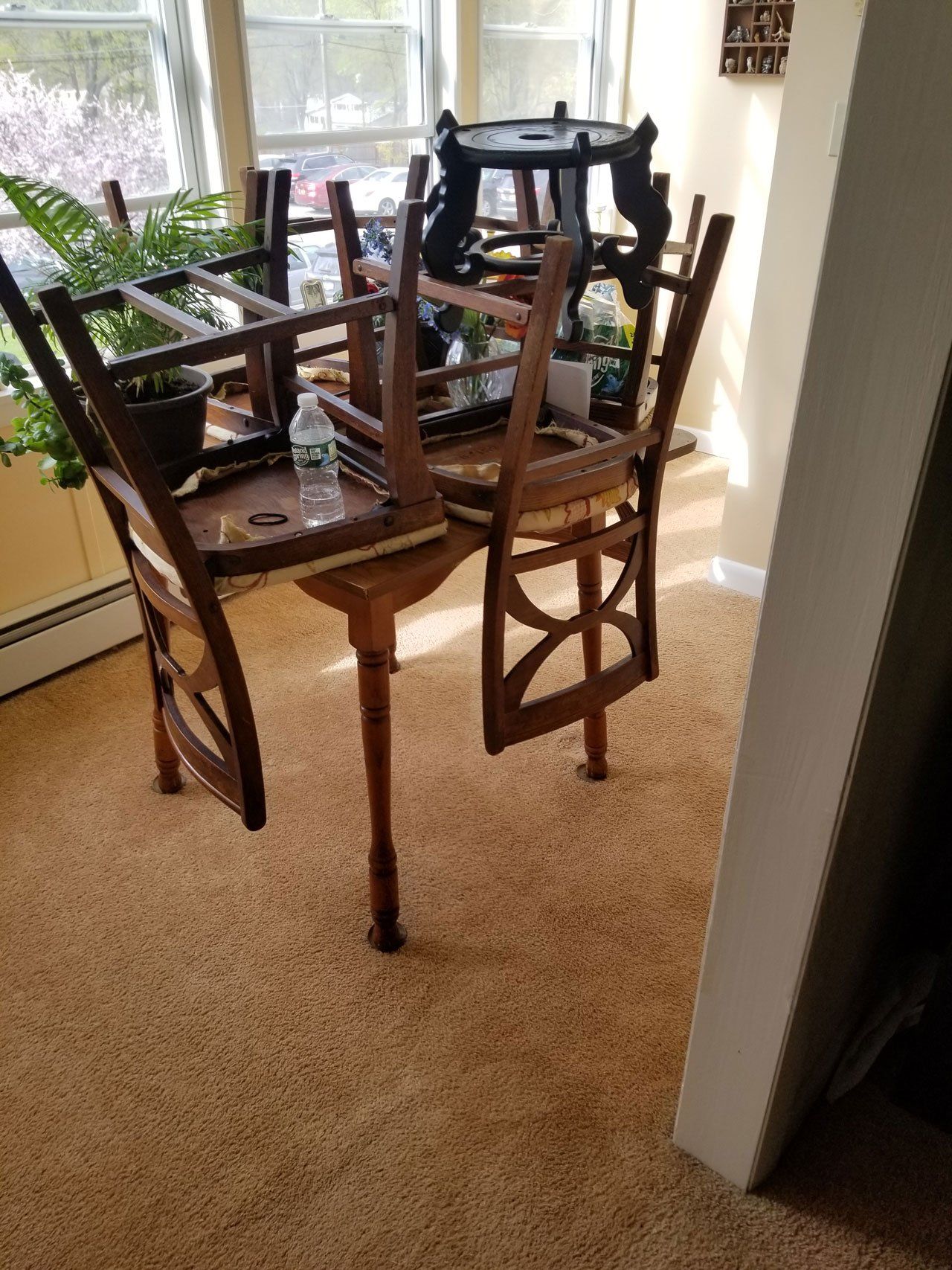 A wooden table in a sunlit room with chairs placed upside down on its surface and a water bottle resting on a seat.