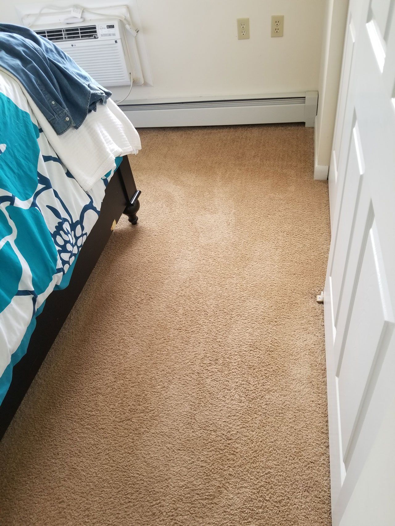 A view of a carpeted bedroom floor next to a bed with a turquoise and white patterned comforter and a white door.