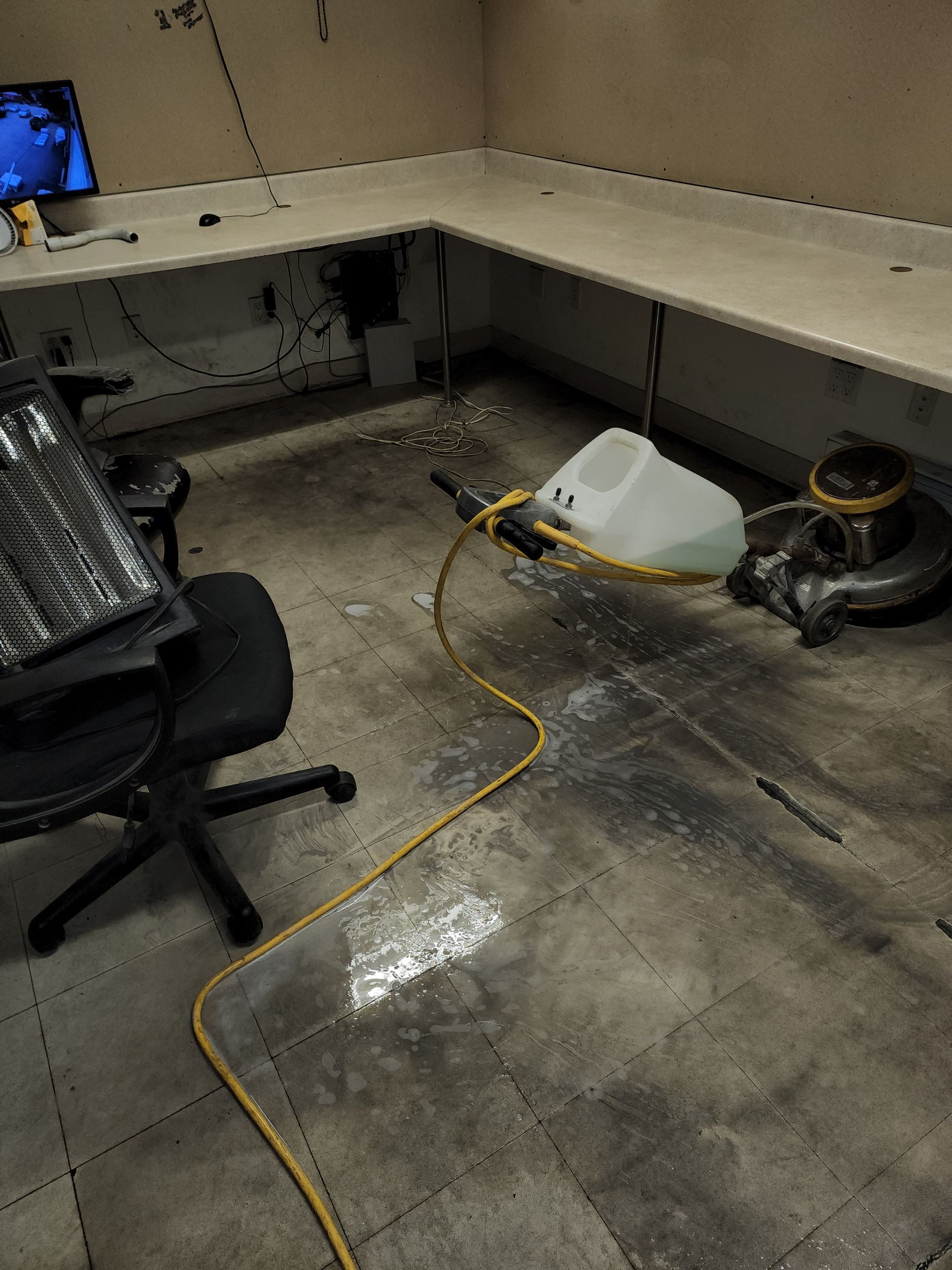 A white floor buffer machine with a liquid tank sits in an office space with a desk chair and a yellow power cord.