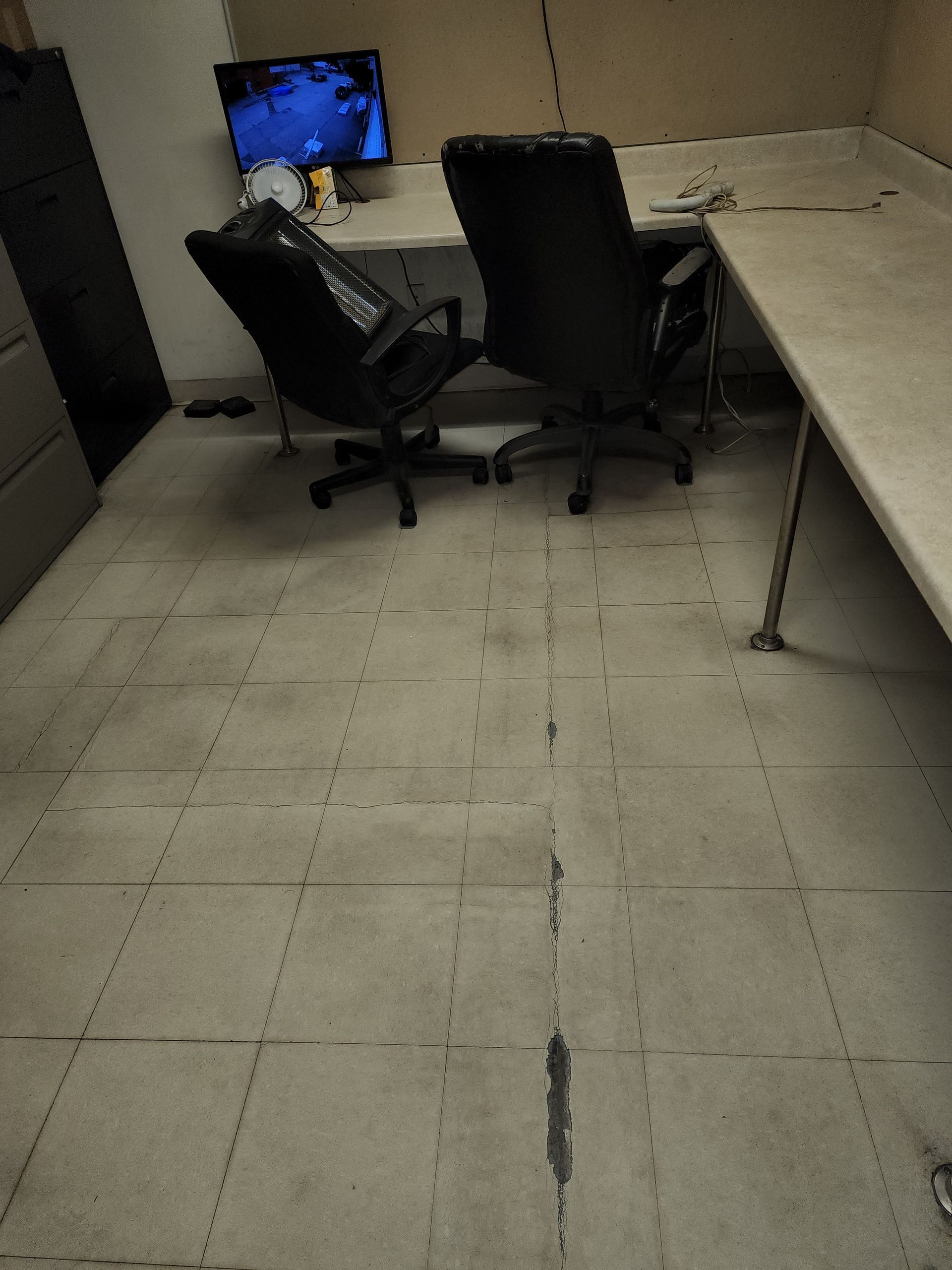 Two black office chairs face a desk in a sparse room with light-colored tile flooring and a computer monitor visible.