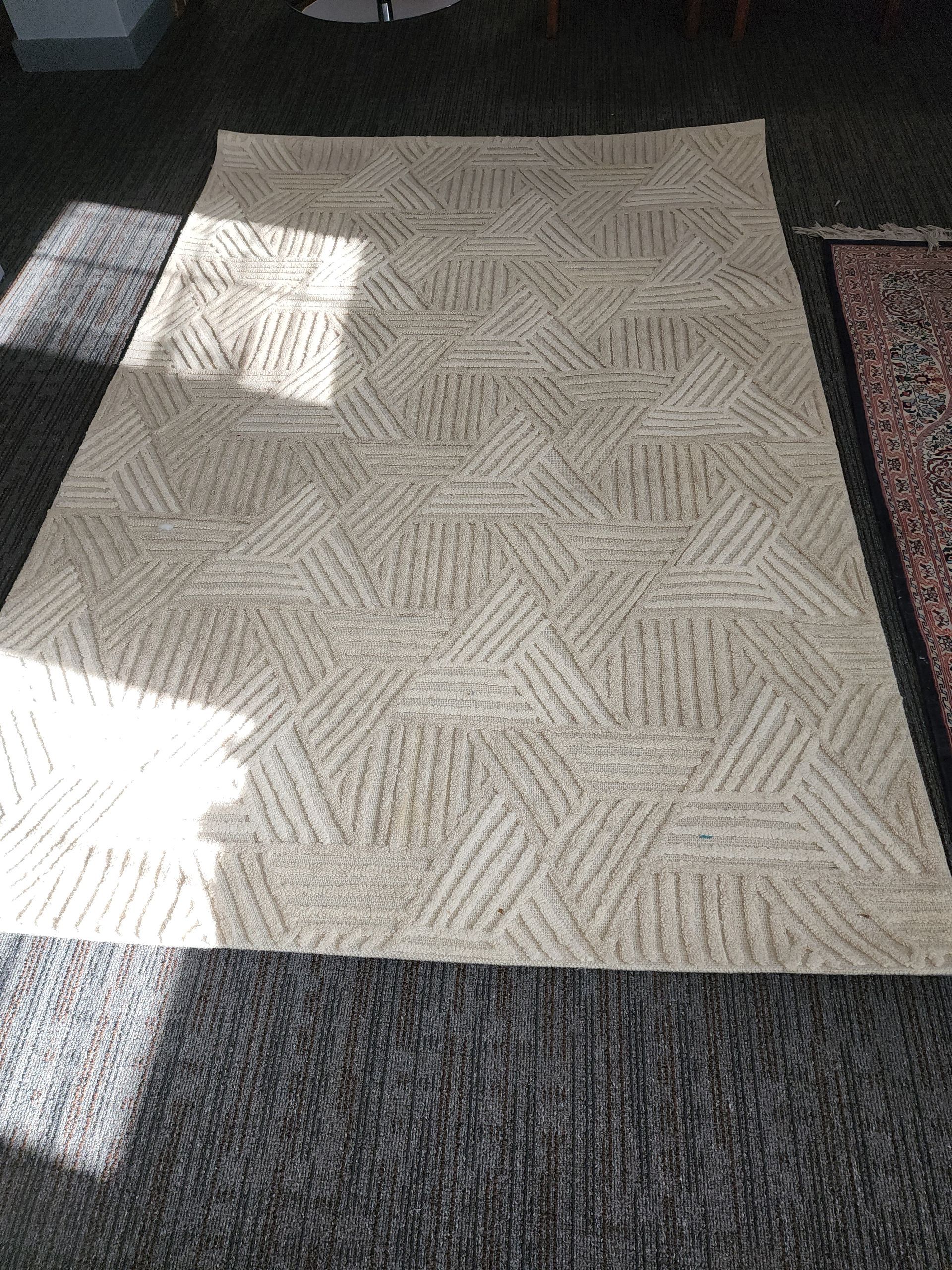 A rectangular area rug with a beige and off-white geometric pattern of intersecting, textured line segments.