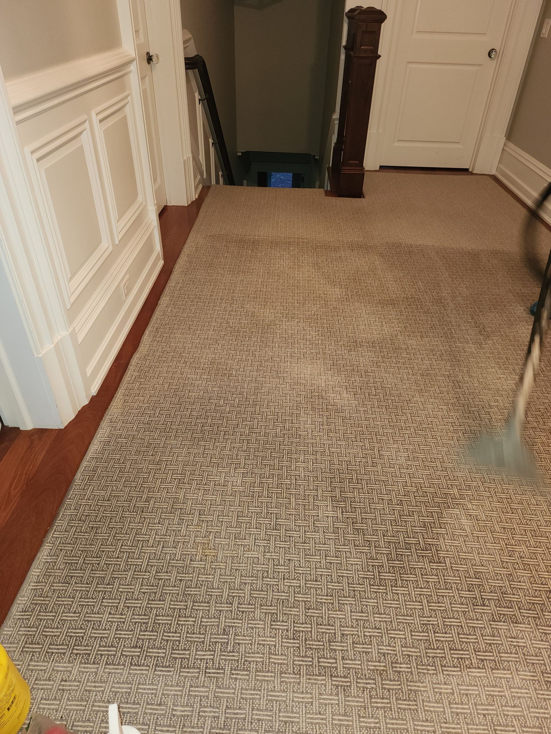 A carpeted hallway with a dark brown banister leads to a staircase; a vacuum attachment rests on the right side.