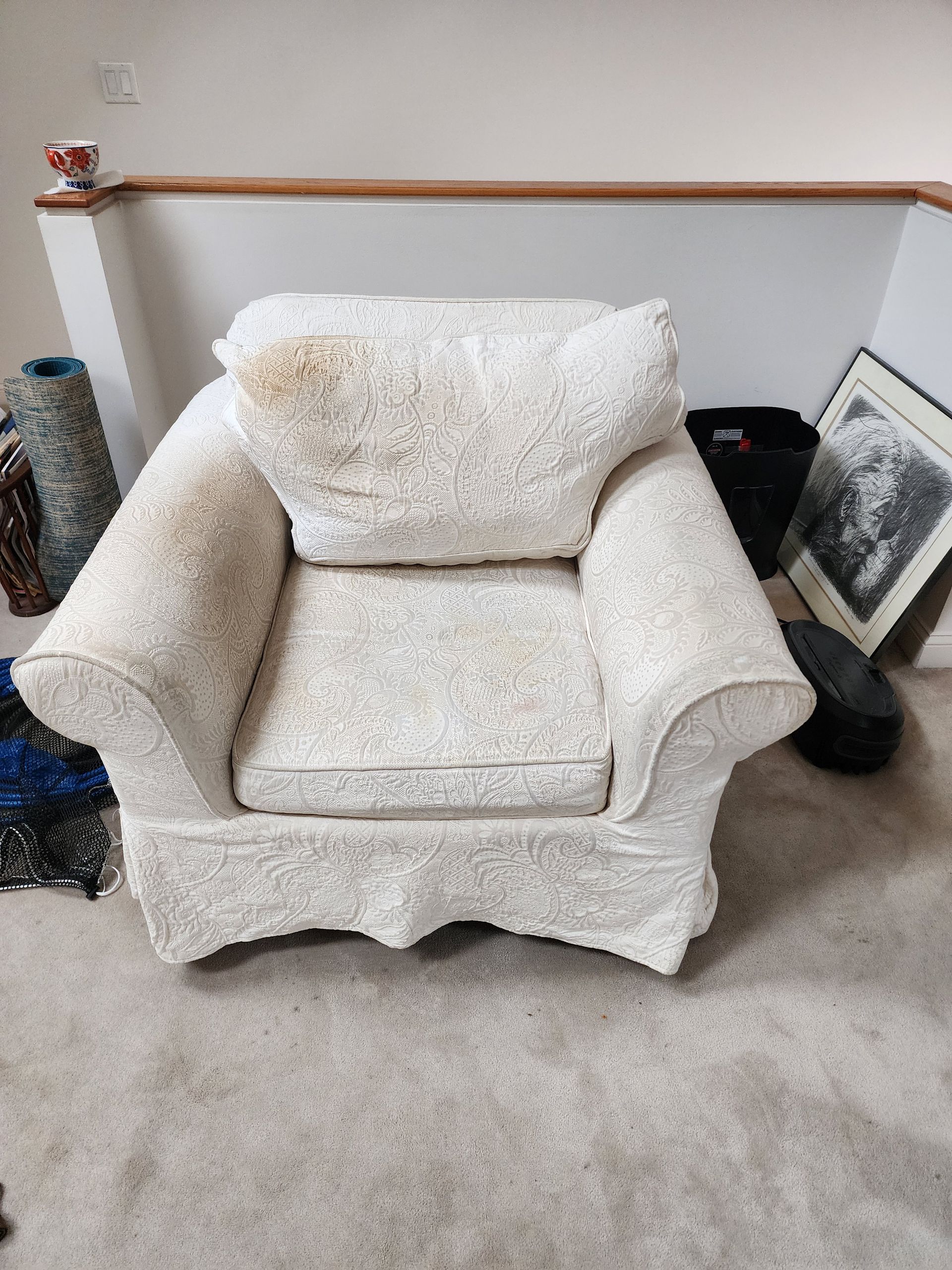 An off-white, patterned upholstered armchair sits on a carpeted floor next to a picture frame and a wastebasket.