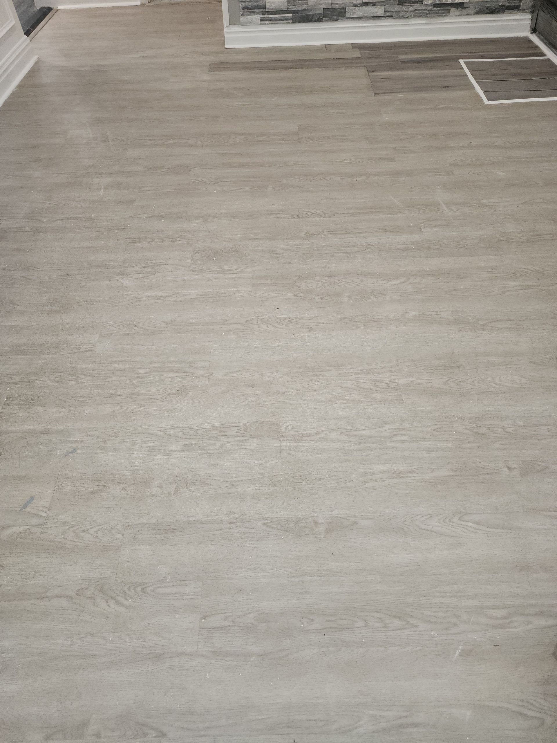 Light grey wood-look laminate flooring panels installed in a room with a white baseboard and a grey stone accent wall.