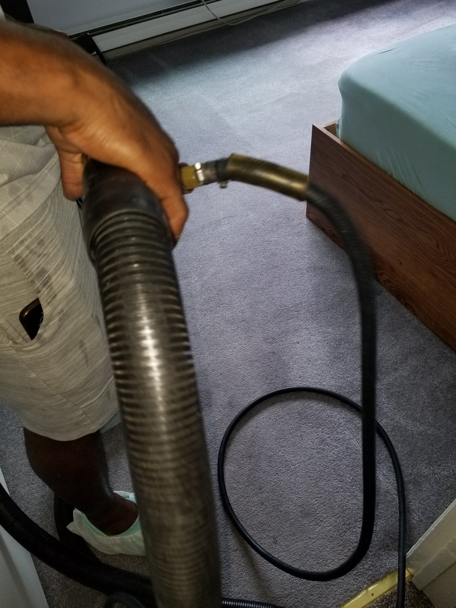 A hand holding a thick vacuum hose with a smaller black tube attached, being used to clean carpeted flooring.