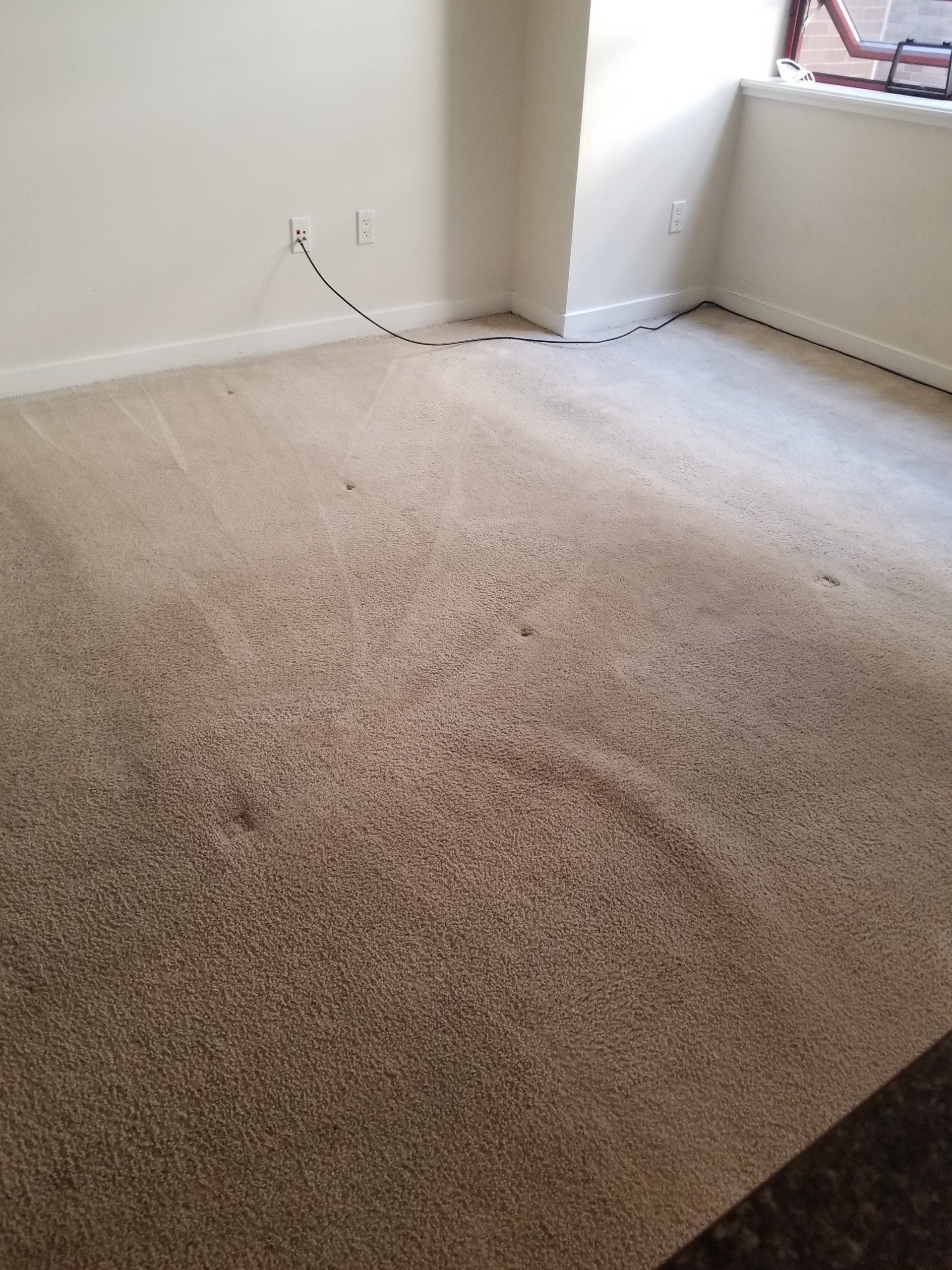 A room with light-colored, worn carpet, white walls, and a visible power cord running along the baseboards.