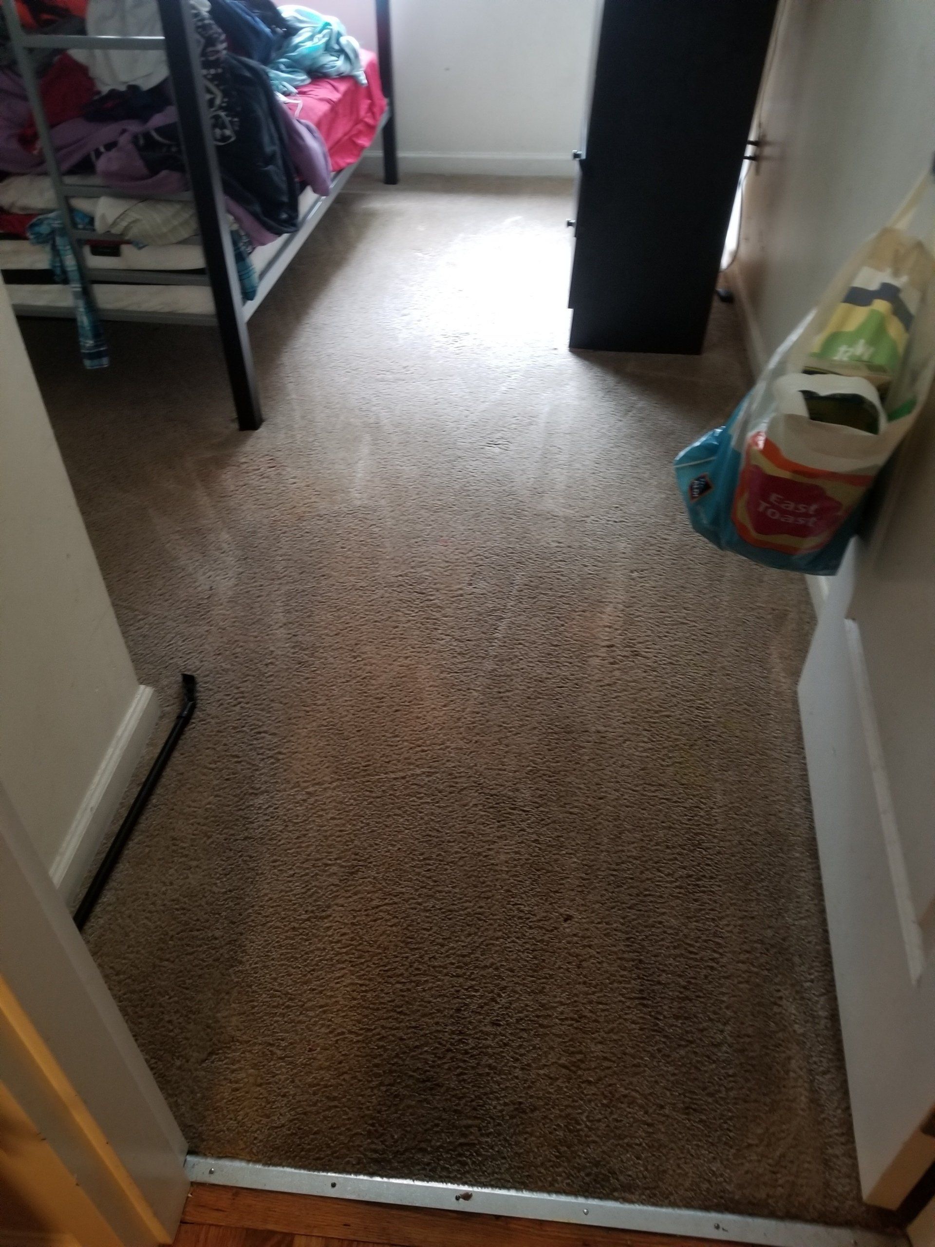 A view from a doorway into a room with brown carpet, a metal bunk bed on the left, and a bag of supplies on the right.