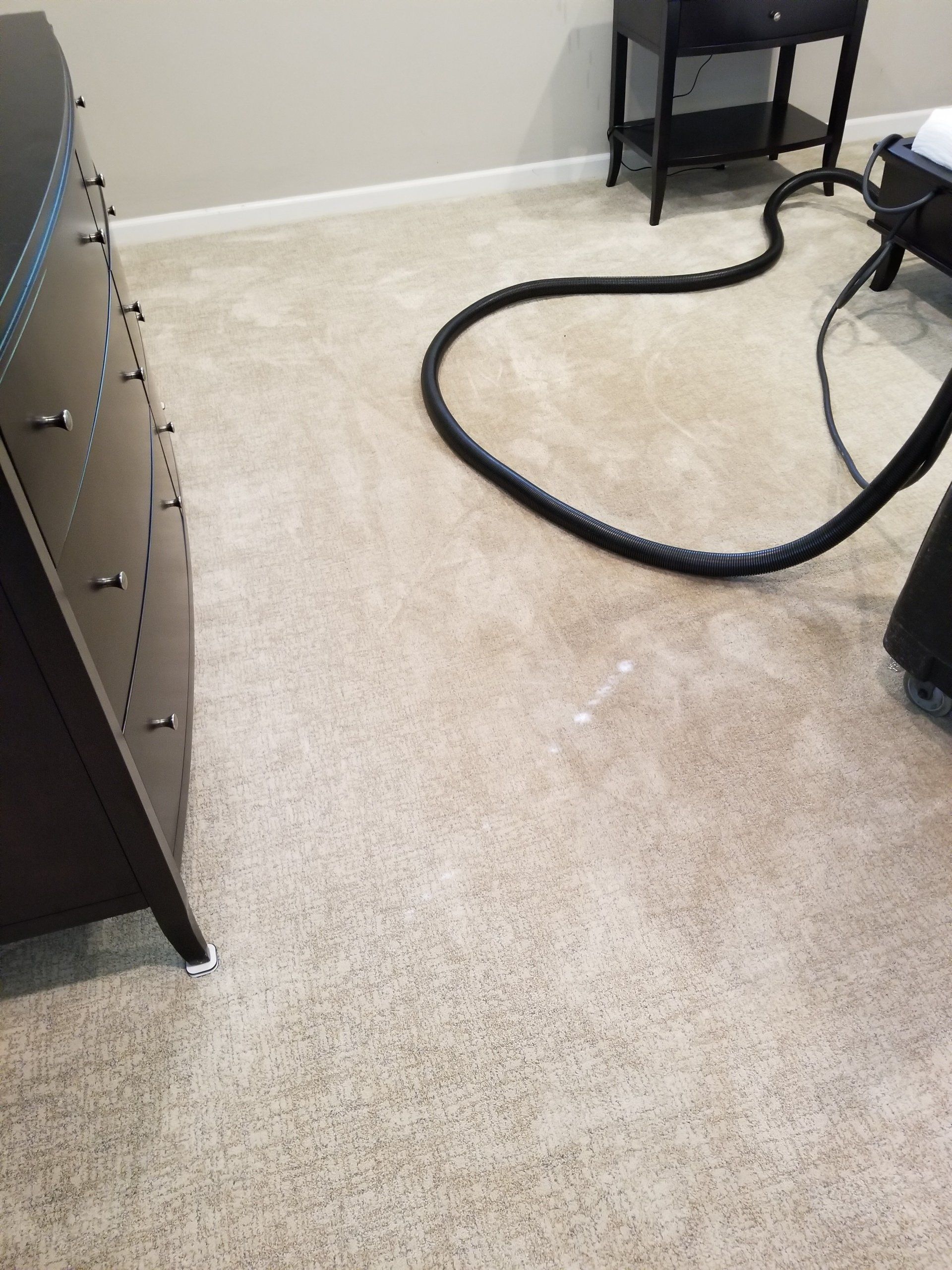 A black vacuum hose lies on a light-colored carpet in a bedroom next to a dark dresser and nightstand.