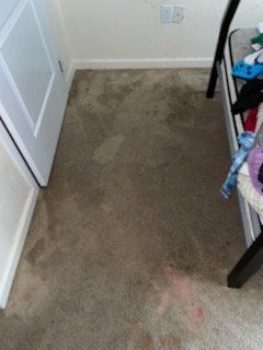 A bedroom carpet showing uneven wear, dirt stains, and a faint red spill near the bottom edge.