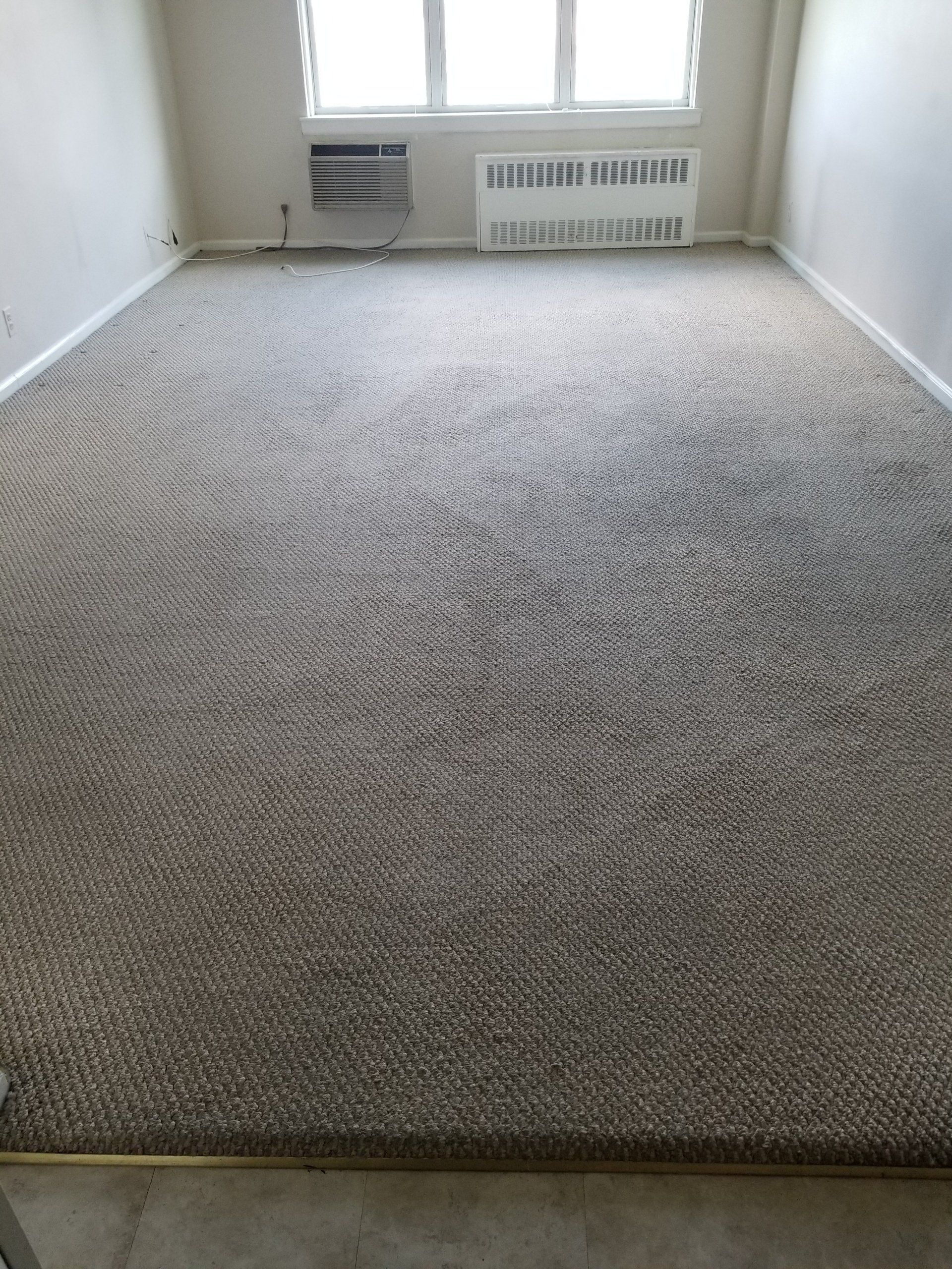 A beige carpeted room with light walls, featuring a window and a wall-mounted heater unit under the sill.