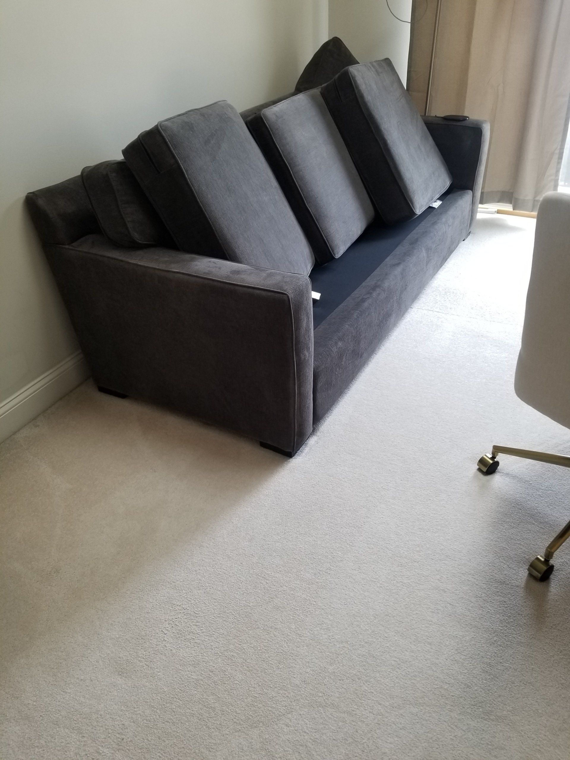A dark grey fabric sofa with three back cushions sits on a light-colored carpet in a room with neutral walls and curtains.