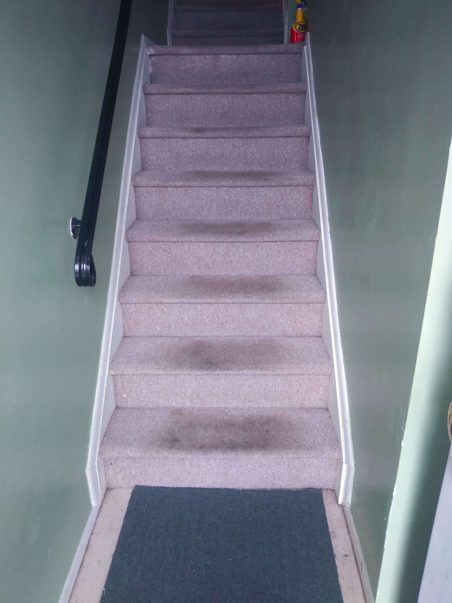 A straight staircase with light beige carpet and a black handrail, leading up between two light green walls.