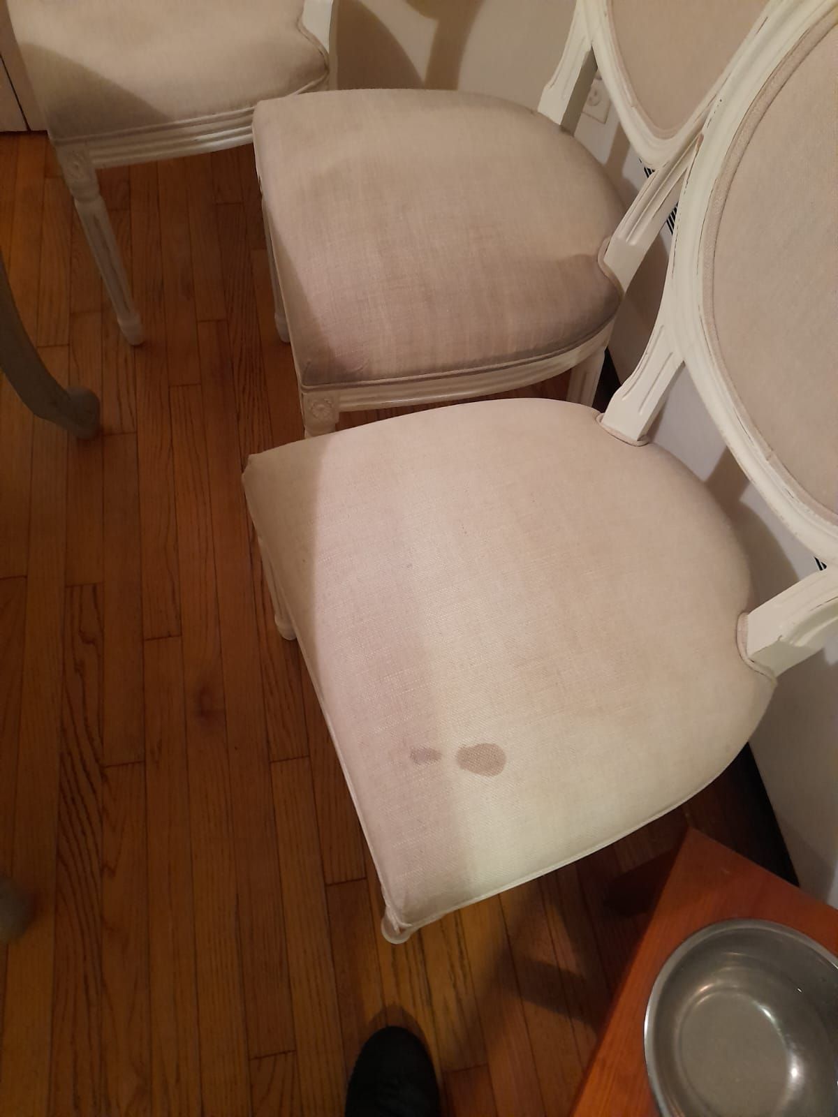 Two off-white upholstered dining chairs on a wooden floor, with a small brown stain on the seat of the front chair.