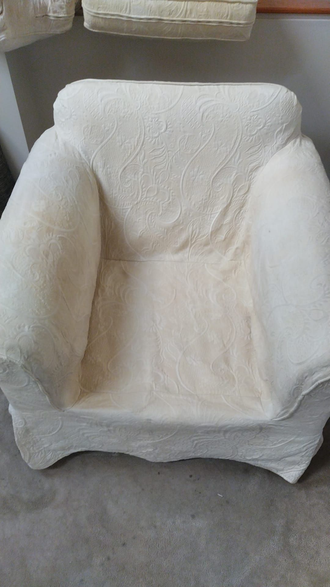 An off-white armchair with a textured, patterned fabric slipcover, shown from a high-angle view on a gray floor.