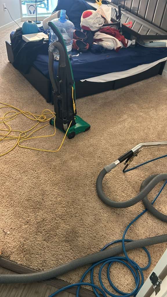 An upright vacuum cleaner and a detached carpet cleaning hose sit on a carpeted floor in a cluttered bedroom.