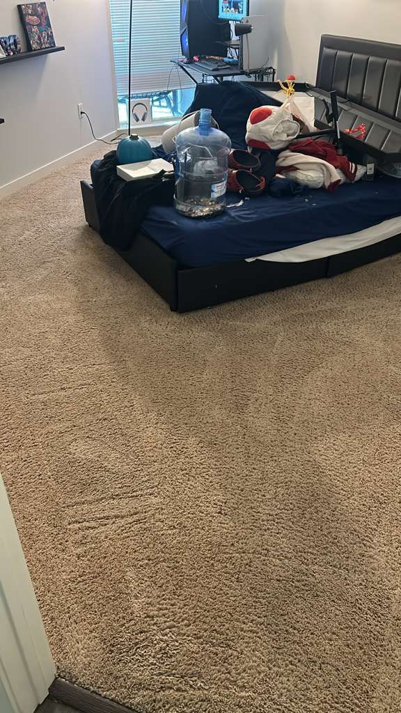 A dark bed frame with a navy blue sheet, clutter, and a large water jug sits on a tan carpeted floor in a bedroom.