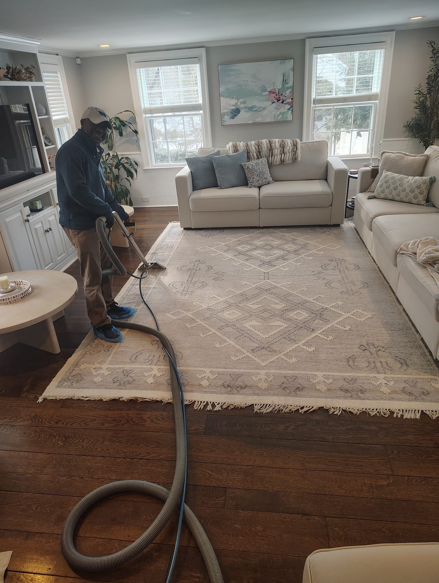 A worker uses a vacuum cleaner wand to clean a large, light-colored patterned rug in a bright, modern living room.