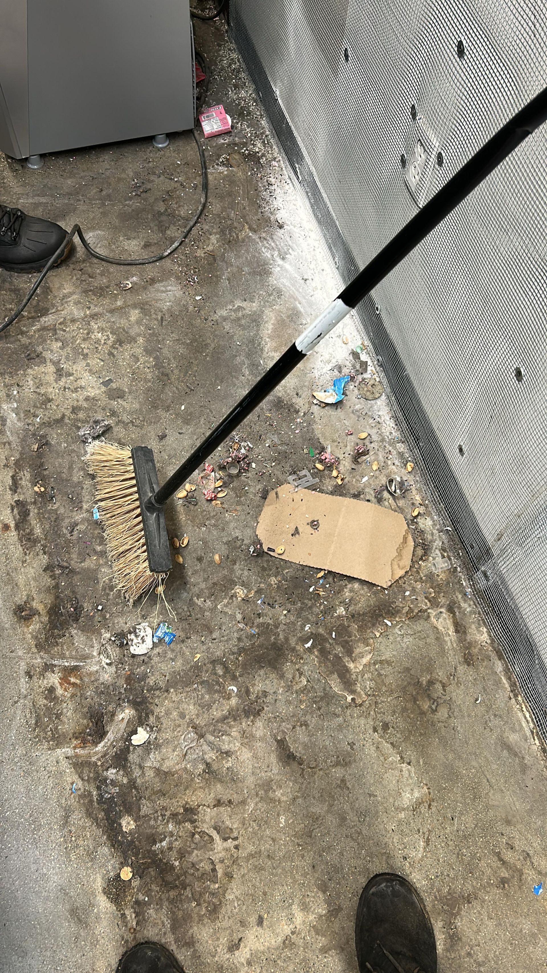 A push broom rests on a dirty concrete floor near a chain-link fence and a piece of discarded cardboard.