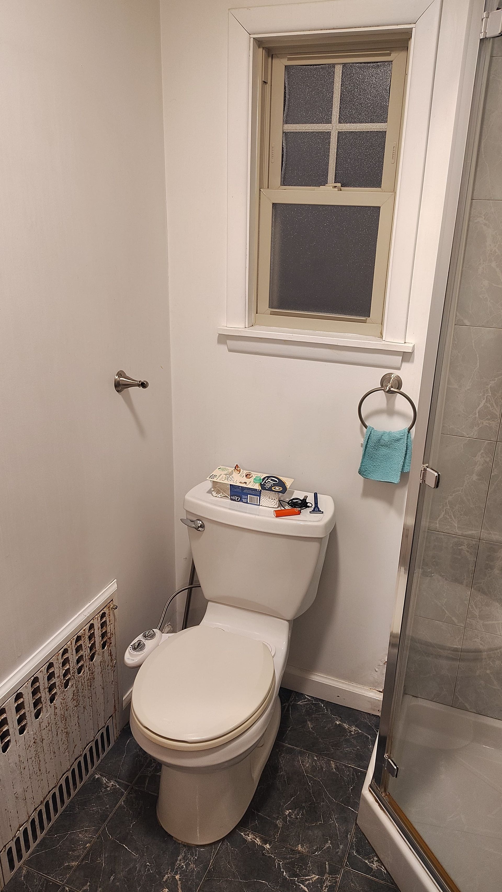 A bathroom with a white toilet featuring a bidet attachment, a small window, a towel ring, and a shower stall on the right.