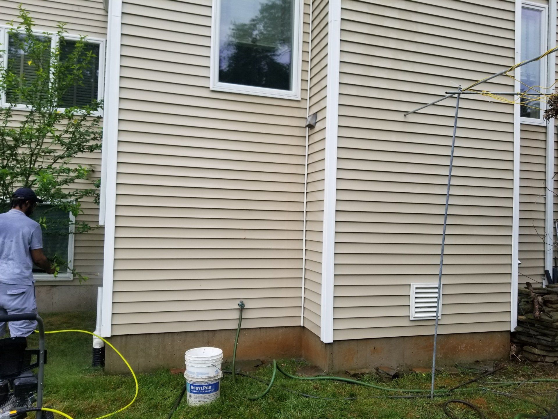 A person washing the beige vinyl siding of a house with a yellow hose, with a white bucket on the grass nearby.