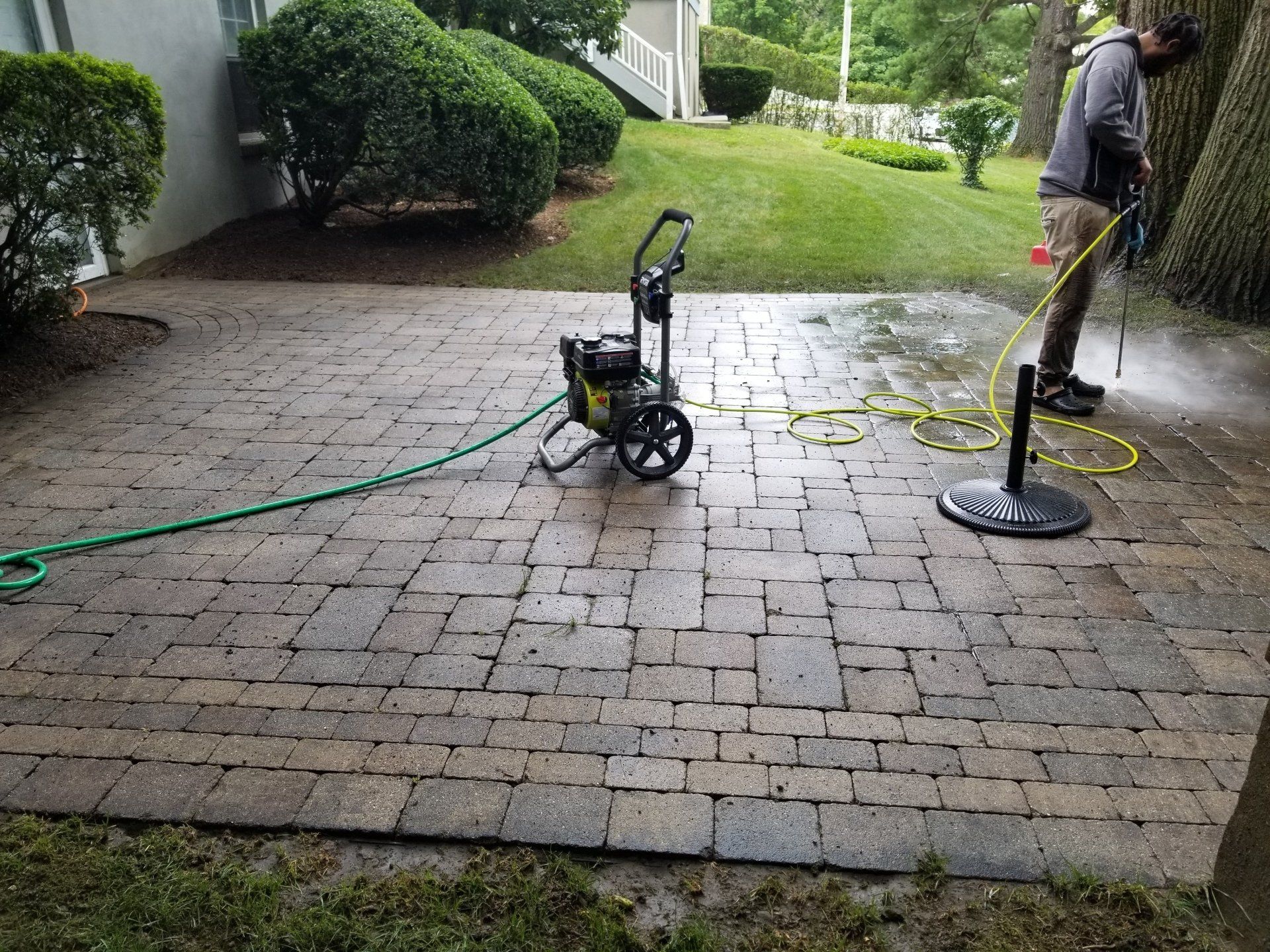 A worker pressure washes a stone patio next to a green lawn, using equipment connected by a yellow hose.